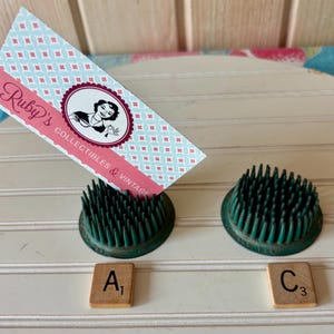 Op de afbeelding: Twee groene bloemenkikkers met metalen pinnen en twee houten Scrabble-tegels met de letters "A" en "C". Een visitekaartje met de tekst "Ruby's Collectibles & Vintage" staat op een van de bloemenkikkers.
