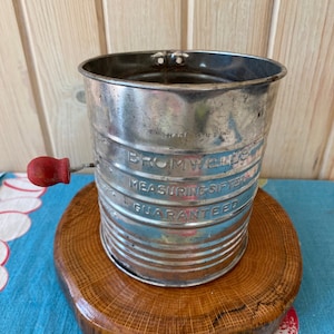 May include: Vintage silver measuring cup with a red handle and the words "BROMWELLS MEASURING SIFTER GUARANTEED" printed on the side. The cup is sitting on a wooden surface.