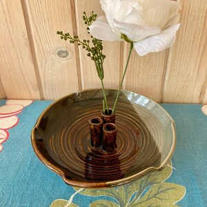 May include: A brown ceramic bowl with a wavy edge and a swirling pattern inside. The bowl has three small brown ceramic cylinders in the centre, which are used as flower vases. A single white flower is arranged in the vases.