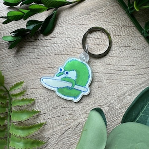 May include: A green worm with two eyes and a toothy grin holding a saw. The worm is on a clear acrylic keychain with a silver metal ring.