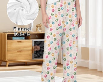 May include: Pajama pants featuring a repeating pattern of multicolored paw prints in shades of pink, yellow, blue, and green on a cream background. An inset circle shows a close-up of the white, soft-textured flannel fabric.