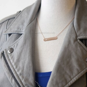 May include: A rose gold necklace with a thin chain and a horizontal bar pendant with a small heart cutout. The necklace is worn by a person wearing a gray leather jacket.
