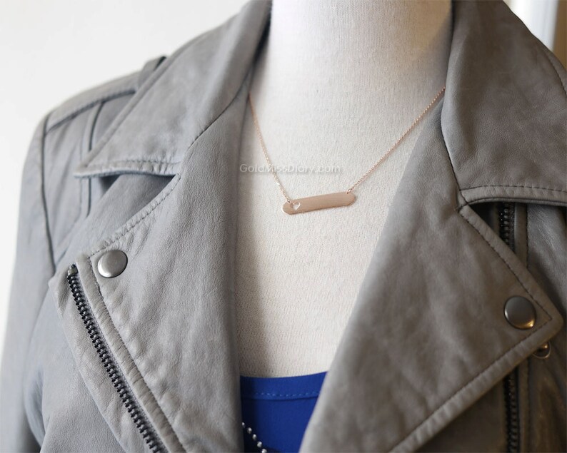 May include: A rose gold necklace with a thin chain and a horizontal bar pendant with a small heart cutout. The necklace is worn by a person wearing a gray leather jacket.