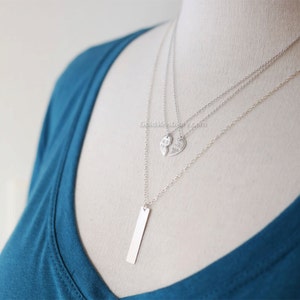 May include: A silver layered necklace with three pendants. The top pendant is a heart shape with the word "best" on one half and "friends" on the other half. The bottom pendant is a long, thin rectangle.