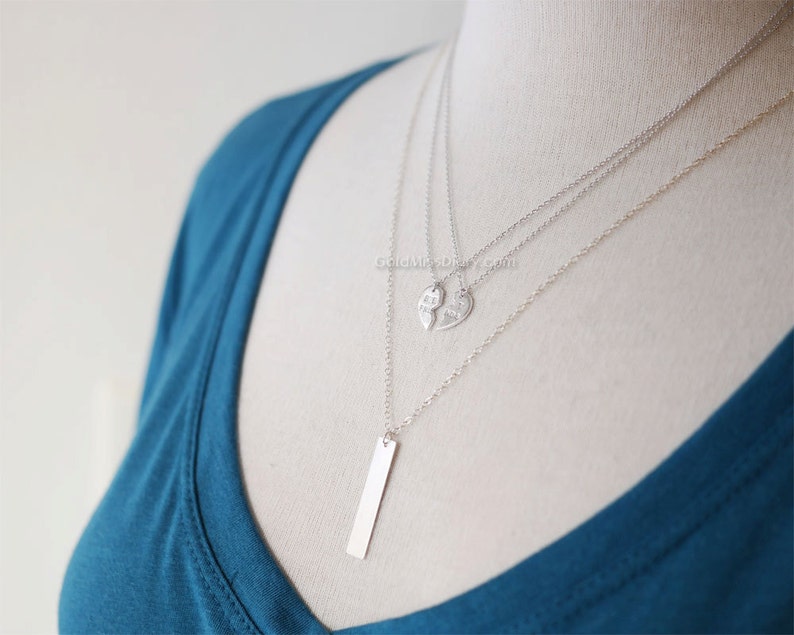 May include: A silver layered necklace with three pendants. The top pendant is a heart shape with the word "best" on one half and "friends" on the other half. The bottom pendant is a long, thin rectangle.