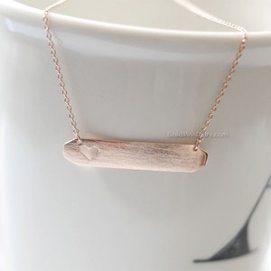 May include: A rose gold necklace with a delicate chain and a bar pendant featuring a heart cutout. The pendant has a brushed finish.