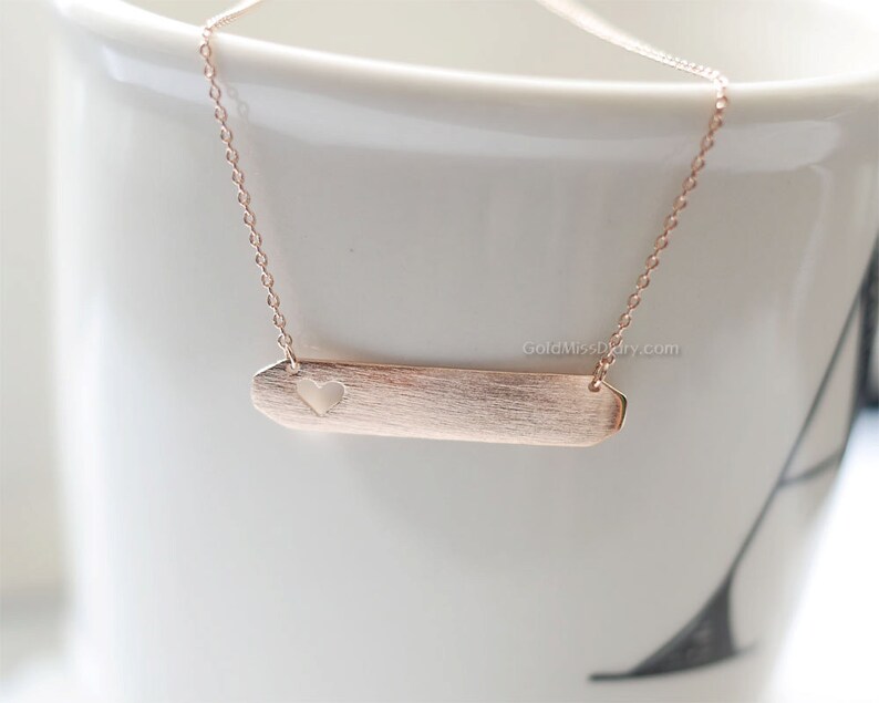 May include: A rose gold necklace with a delicate chain and a bar pendant featuring a heart cutout. The pendant has a brushed finish.
