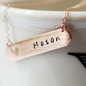 May include: A rose gold necklace with a bar pendant that has the name "Mason" engraved on it. The bar pendant has a small heart cut-out.