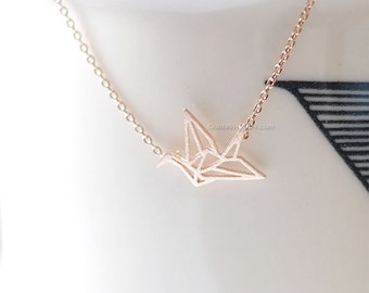 Peut inclure: Collier avec un pendentif grue origami en or rose. Le pendentif géométrique est suspendu à une fine chaîne. La grue présente des plis et des angles visibles. Le collier est présenté sur une surface blanche.