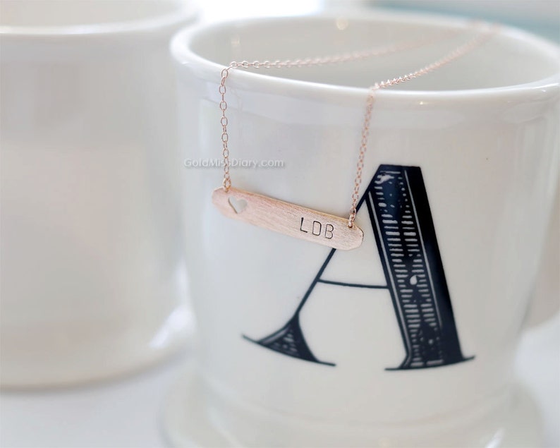 May include: A rose gold necklace with a heart cutout and a bar pendant engraved with the initials "LDB".