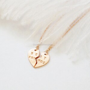 May include: Two gold necklaces with matching heart pendants that say "Best Friends" on them. The necklaces are on a white feathery background.