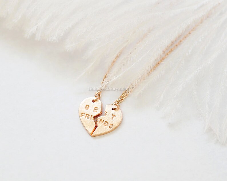 May include: Two gold necklaces with matching heart pendants that say "Best Friends" on them. The necklaces are on a white feathery background.