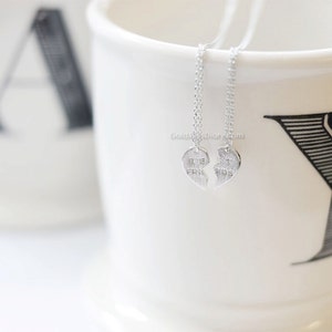 May include: Two silver necklaces with heart pendants that say "Best Friends" on them. The necklaces are hanging from a white mug with black lettering.