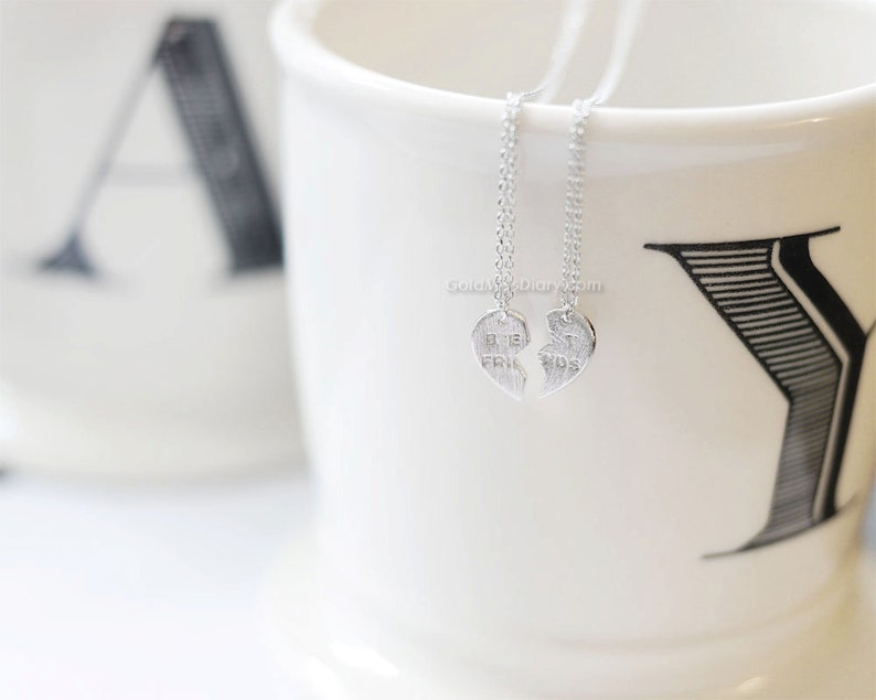 May include: Two silver necklaces with heart pendants that say "Best Friends" on them. The necklaces are hanging from a white mug with black lettering.