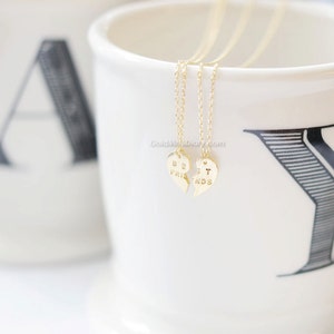 May include: Two gold necklaces with matching heart pendants that say "Best Friends" on them. The necklaces are displayed on a white mug with black lettering.