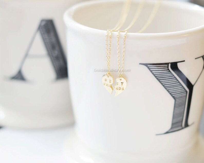 May include: Two gold necklaces with matching heart pendants that say "Best Friends" on them. The necklaces are displayed on a white mug with black lettering.