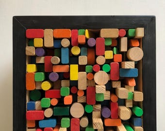 May include: A colorful wooden art piece featuring an array of geometric shapes in various colors, including red, orange, yellow, green, blue, and purple. The shapes are arranged within a black wooden frame, creating a textured, abstract design.