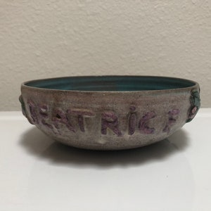 May include: A gray ceramic bowl with a teal glaze inside. The bowl has the word "PATRIC" written in purple on the outside.
