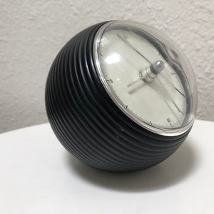 May include: A black, spherical clock with a ribbed texture and a clear, domed cover. The clock face is white with black numerals and hands. The clock is approximately 5 inches in diameter.