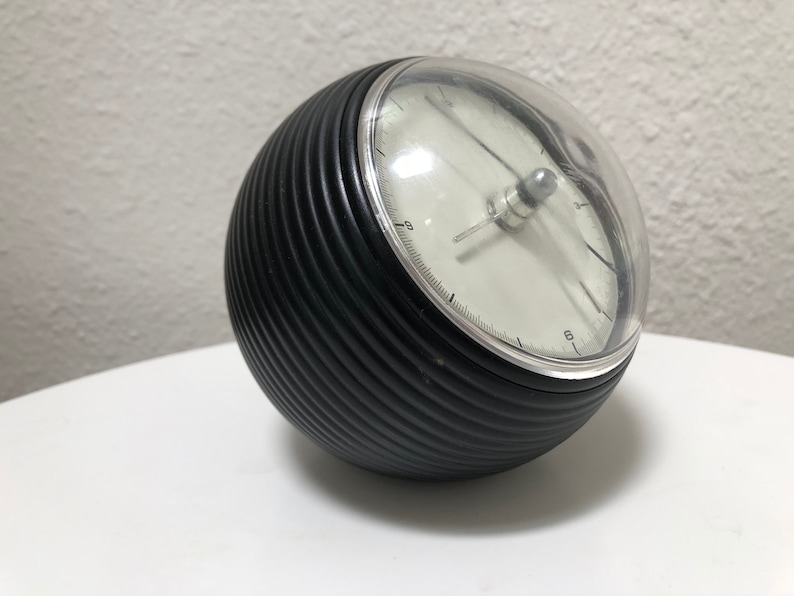 May include: A black, spherical clock with a ribbed texture and a clear, domed cover. The clock face is white with black numerals and hands. The clock is approximately 5 inches in diameter.
