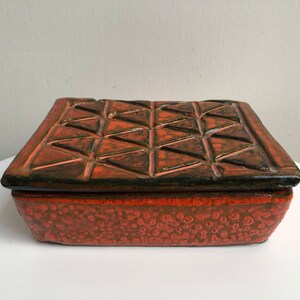 May include: A ceramic box with a geometric pattern on the lid. The lid is dark brown with a red glaze, and the base is a solid red with a speckled glaze.