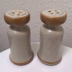 May include: A pair of ceramic salt and pepper shakers with brown tops and a speckled white finish. The tops have a decorative design.