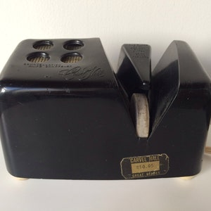 May include: Black electric knife sharpener with a white sharpening stone and a gold label that reads "Carvel Hall $10.95 Great Berries".