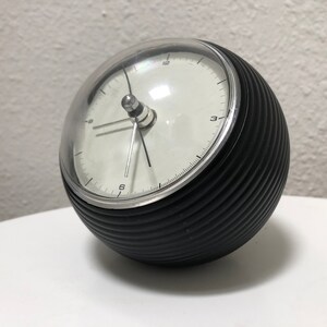 May include: A black and silver spherical alarm clock with a clear, domed top. The clock face is white with black numerals and hands. The clock's body has a ribbed texture.