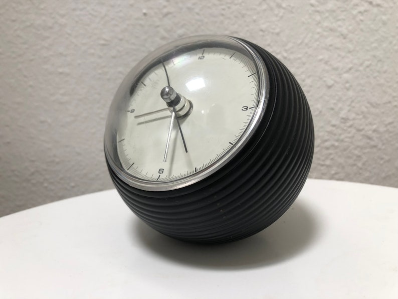 May include: A black and silver spherical alarm clock with a clear, domed top. The clock face is white with black numerals and hands. The clock's body has a ribbed texture.