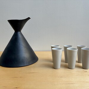 May include: A black ceramic sake carafe with a conical base and a spout, paired with six off-white ceramic shot glasses. The objects are arranged on a light wooden surface.