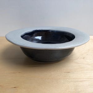 May include: A small, white-rimmed ceramic bowl with a dark blue glaze. The bowl is sitting on a light wood surface.