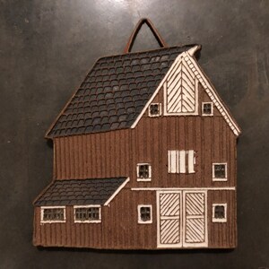 May include: A brown ceramic barn wall hanging with white accents. The barn has a gabled roof, windows, and a double door. The barn has a hanging loop at the top.