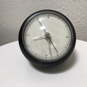 May include: A black and silver analog clock with a white face. The clock has a round, ribbed black casing and silver hands. The clock face displays the numbers 1 through 12. The clock is sitting on a white surface.