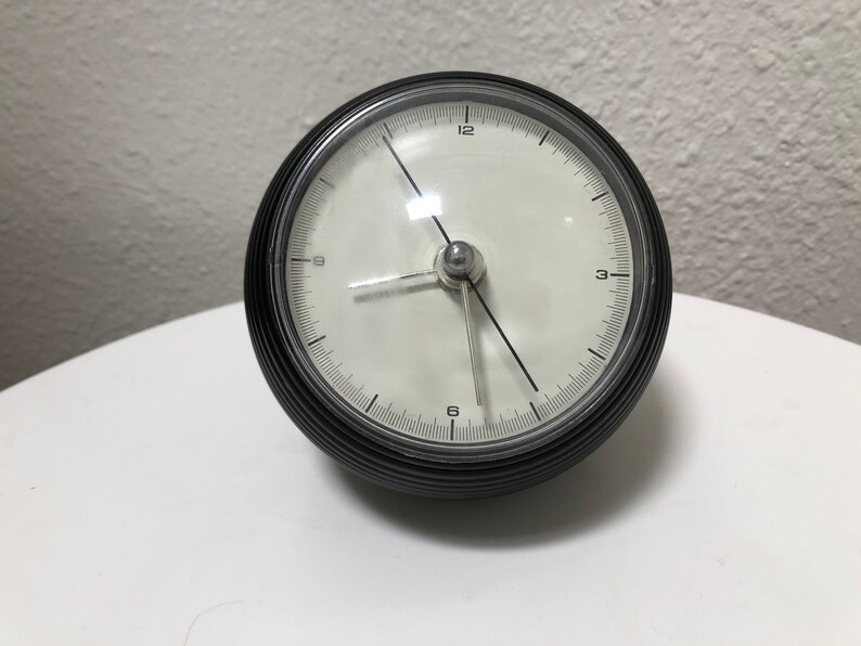 May include: A black and silver analog clock with a white face. The clock has a round, ribbed black casing and silver hands. The clock face displays the numbers 1 through 12. The clock is sitting on a white surface.