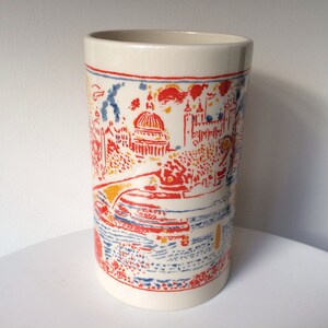 May include: White ceramic mug with a colorful illustration of a city skyline. The illustration features red, yellow, and blue buildings and a river.