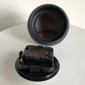 May include: A disassembled black clock with a round, ribbed body and a separate clock mechanism. The clock mechanism is black and clear, showing the internal gears. The clock's interior is a dark brown color.