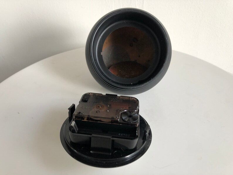 May include: A disassembled black clock with a round, ribbed body and a separate clock mechanism. The clock mechanism is black and clear, showing the internal gears. The clock's interior is a dark brown color.