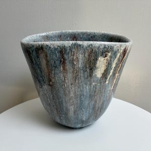 May include: A wide-mouthed ceramic vessel with a tapered base, featuring a mottled glaze in shades of slate blue, grey, and earthy brown. The surface displays vertical drip patterns and textured, light-colored patches throughout the exterior.