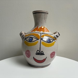 May include: A ceramic vase with a painted face design. The vase features a light gray base with a smiling face, blue eyes, and orange and yellow accents. It has two small handles and a narrow neck.