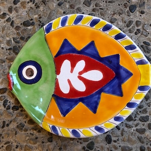 May include: A hand-painted ceramic fish-shaped plate. The plate features a green head, an orange body with a red and white design, and blue and yellow accents. The plate is approximately 10 inches long.