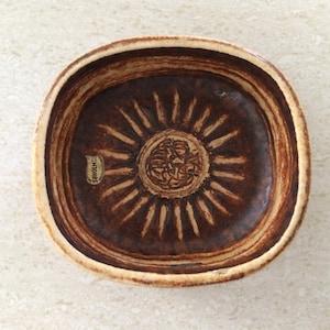 May include: A square, brown ceramic bowl with a sunburst design in the center. The bowl has a light beige rim and a dark brown interior. The center of the bowl features a detailed, embossed design. The label reads "Soholm Bornholm".