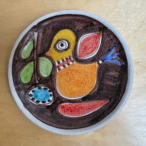 May include: A hand-painted ceramic plate with a whimsical bird design. The bird is orange with yellow head, red and blue accents, and a black and white striped neck. The plate has a brown background and a white rim.