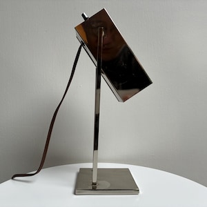 May include: A modern, silver-toned desk lamp with a rectangular shade and a square base. The lamp has a slender, vertical support and a brown power cord. The shade is angled, and the lamp is on a white surface.