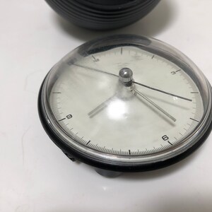 May include: A small, round clock with a clear, domed cover. The clock face is white with black numerals and hands. The clock's bezel is black, and the clock is sitting on a white surface. A black, ridged object is in the background.