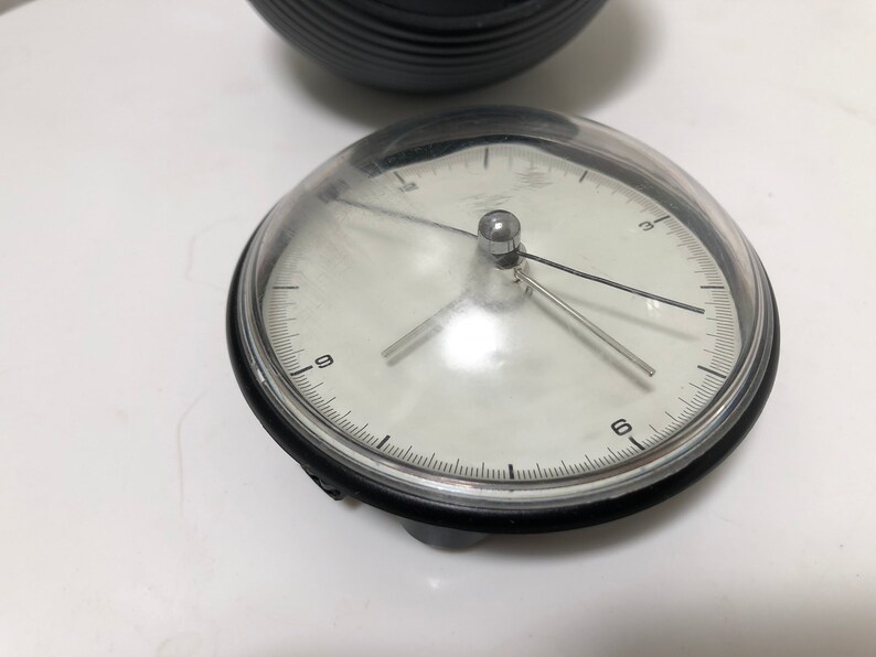 May include: A small, round clock with a clear, domed cover. The clock face is white with black numerals and hands. The clock's bezel is black, and the clock is sitting on a white surface. A black, ridged object is in the background.