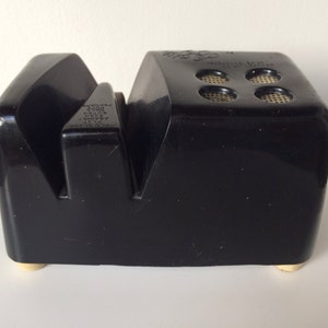 May include: Black plastic knife sharpener with a textured surface and gold feet. The sharpener has a small hole on the top with the text "Model No. 100" printed on it.