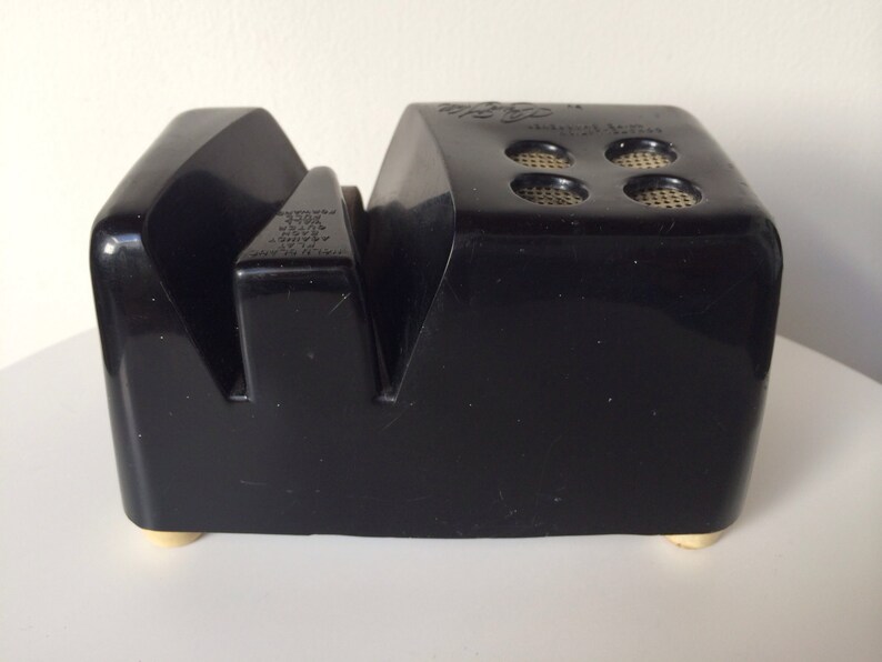 May include: Black plastic knife sharpener with a textured surface and gold feet. The sharpener has a small hole on the top with the text "Model No. 100" printed on it.
