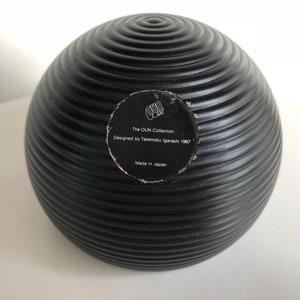 May include: A black, spherical decorative object with a ridged surface. The object has a circular sticker with text that reads "The OUN Collection, Designed by Takenobu Igarashi 1987, Made in Japan."