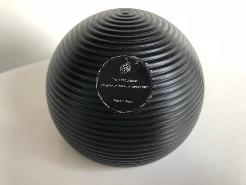 May include: A black, spherical decorative object with a ridged surface. The object has a circular sticker with text that reads "The OUN Collection, Designed by Takenobu Igarashi 1987, Made in Japan."