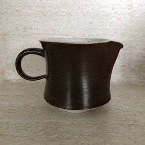May include: A brown ceramic creamer with a white rim and a handle. The creamer has a slightly flared shape and a smooth, glossy finish.